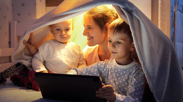 Улыбающаяся Мать Двумя Мальчиками Сидящими Одеяле Постели Пользующимися Планшетным Компьютером — стоковое фото