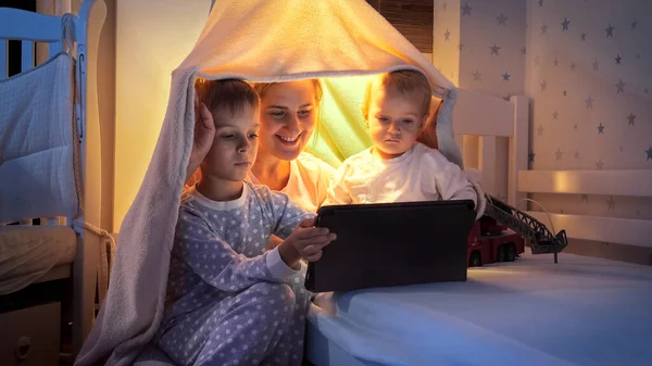年轻的母亲晚上和两个儿子在床上看平板电脑上的视频 有时间在一起的家庭 带着小玩艺儿的孩子 快乐的童年和娱乐 — 图库照片