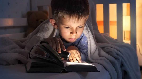 パジャマ姿のかわいい男の子がベッドで懐中電灯で本を読んでいる 子供の教育 — ストック写真
