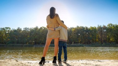 Ormandaki gölde oğlunu kucaklayan ve öpen genç bir anne. Aile açık havada, mutlu ebeveynlik ve çocukluk, sonbahar manzarası, çocuklar ve ebeveynler dışarıda dinleniyor.