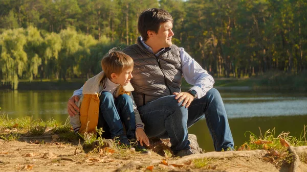 幸せな父親は彼の息子と川岸に座って日没を見ている 屋外の家族 幸せな子育てと子供時代 秋の風景 子供と親外でリラックス — ストック写真