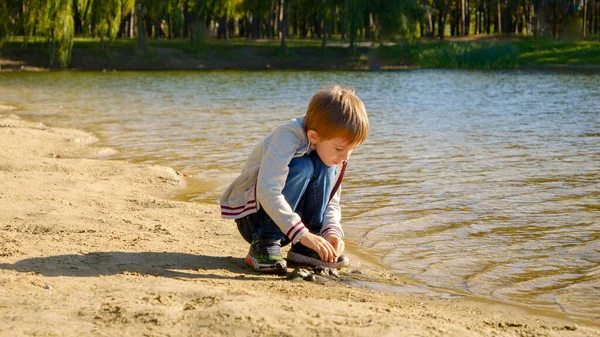 小男孩坐在河岸上玩湿沙子 孩子们在户外玩耍 孩子们在海滩上 湖在城市公园 — 图库照片