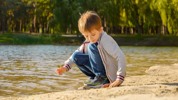 Szczęśliwy Wesoły Chłopiec Siedzący Nad Jeziorem Wrzucający Mokry Piasek Wody — Zdjęcie stockowe