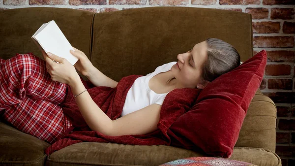 リビングルームでソファで本を読んでいる美しい若い女性 女性のレジャーで休んでいる人々 — ストック写真