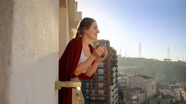 美しい女性がコーヒーを飲みながらバルコニーに立ち 朝の街の景色を眺める — ストック写真