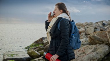 Deniz kenarında dinlenen genç bir kadın, elinde sıcak bir çay termosuyla vahşi ve engebeli kıyı şeridine hayran. Turizm, seyahat veya yaşam tarzı içeriğinde kullanım için ideal