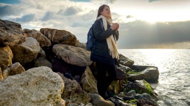 Genç, gülümseyen bir kadın bir fincan sıcak çayla uçurumlarda duruyor ve soğuk deniz dalgalarını seyrediyor..