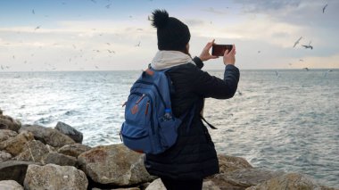 Kışlık elbiseli bir kadın, deniz kenarındaki kayalıklarda dikilmiş, akıllı telefonuyla kıyıya vuran soğuk ve fırtınalı dalgaların fotoğraflarını çekiyor. Seyahat, turizm ve macera