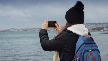 Kumsalda fırtınalı bir kış gününün güzelliğini yakalayan genç bir kadın. Arkasındaki kayalara çarpan akıllı telefonuyla. Seyahat, macera ve keşif.