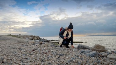 Genç bayan yürüyüşçü kayalık deniz sahillerinde yürüyor ve soğuk rüzgarlı bir günde deniz kabukları ve çakıl taşları topluyor. Yürüyüş, seyahat, keşif, turizm ve seyahat kavramı