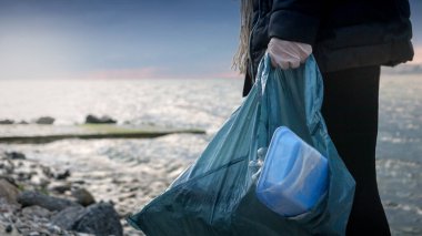 Sahilden topladığı çöplerle dolu plastik bir poşeti tutan bir kadının yakın çekimi. Çevre koruma kavramı, sürdürülebilirlik ve çevre dostu alışkanlıklar.