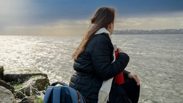 長い髪をした若い女性が海岸の大きな岩の上に座って 海の上に嵐の天気を見ています 冬の観光 休暇の概念 — ストック写真