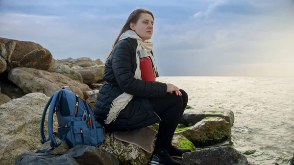 年轻的女人留着长长的飘扬的头发 坐在岩石的海岸线上 手里拿着热茶 凝视着外面汹涌的大海 最适合旅行或度假 — 图库照片