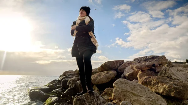 年轻的女人站在悬崖上 看着海面上冬日的落日 肩上背着背包 探险和旅行的概念 — 图库照片