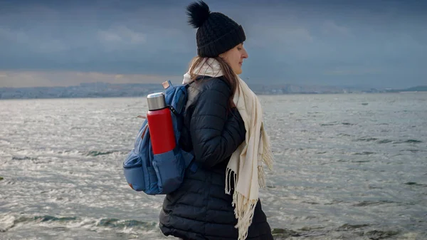 バックパックと魔法瓶を装備した女性ハイカーは 寒い曇りの日に岩の多い海のビーチを探索します 冒険をテーマにしたプロジェクトに加えて — ストック写真