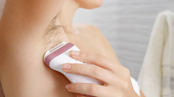 Closeup of young woman with long armpit hair making depilation with electric epilator. Concept of hygiene, natural beauty, feminity and body hair growth