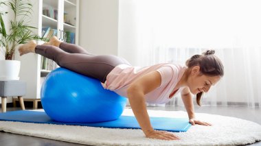 Smiling young woman doing exercises and balancing on fitball at living room. Concept of healthcare, sports and yoga at home clipart
