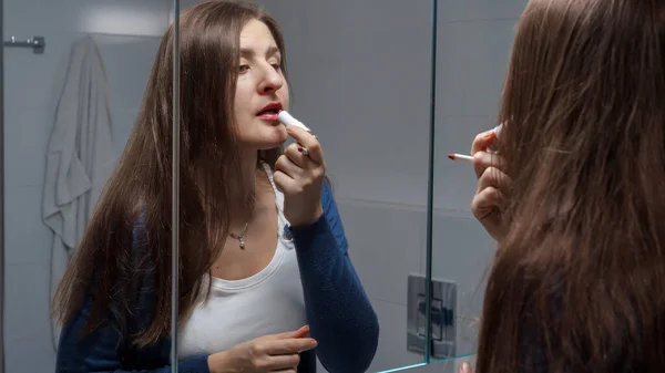 Young brunette woman looking in mirror and painting lips with red lipstick.