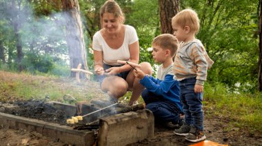 Mutlu bir anne ve iki çocuk orman kampında şenlik ateşinde sosis pişiriyorlar. Aktif eğlence, kamp yapan çocuklar, doğada aile tatili.