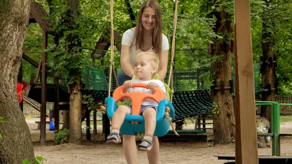 Uśmiechnięta Kobieta Popycha Synka Huśtawce Placu Zabaw Parku Dzieci Bawiące — Zdjęcie stockowe