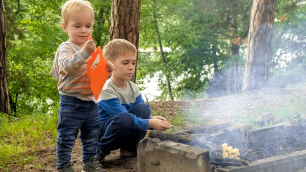 两个小男孩坐在森林篝火边 用木棍烤面包 积极的闲暇 露营中的儿童 自然的家庭假期 — 图库照片