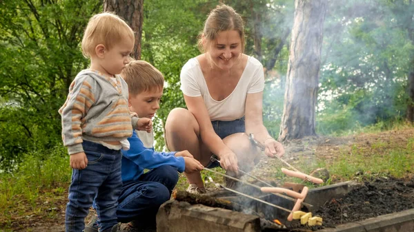 Δύο Αγόρια Κοιτάζουν Μητέρα Μαγειρεύει Λουκάνικα Στη Φωτιά Στο Δάσος — Φωτογραφία Αρχείου