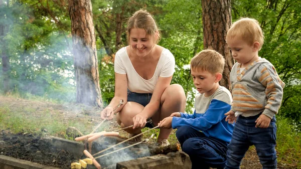 有两个儿子的年轻母亲在森林里用篝火烤烤肉和烤香肠 — 图库照片