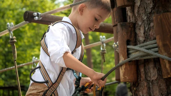 ロープアドベンチャーパークに登りながら 小さな男の子は木に安全ロープフックをクランプします アクティブな子供時代 健康的なライフスタイル 屋外で遊んでいる子供 自然の中で子供 — ストック写真