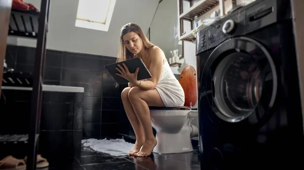 A woman multitasking, sitting on a toilet, and working on a tablet computer. Importance of being efficient and productive in all situations