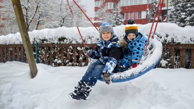 Kış eğlencesi ve kahkahalar iki kardeş karla kaplı bir parkta sallanırken. Açık hava aktivitesi ve aile eğlencesi