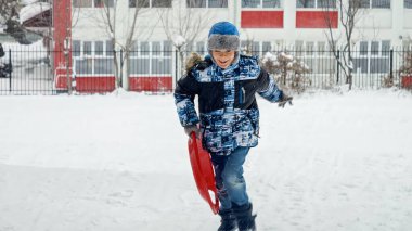 Çocuk mutlu bir şekilde karda plastik kızaklarla koşuyor, temiz havanın ve kış manzarasının tadını çıkarıyor.