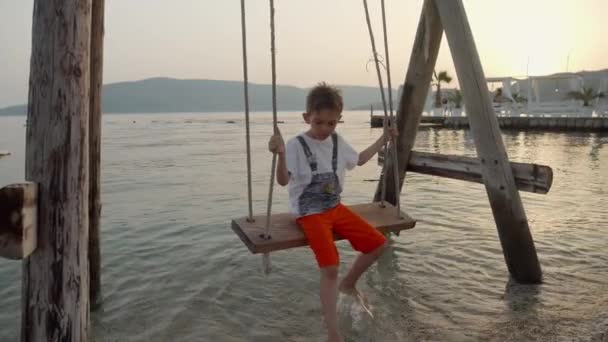 木製のロープでスイングする小さな男の子のゆっくりとした動きは 日没時に海のビーチで泳ぎます 夏休み 観光について — ストック動画