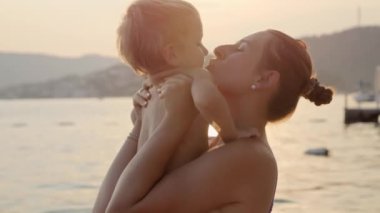 Mayolu mutlu bir anne ve gün batımında deniz sahilinde sarılıp eğlenen bir bebek. Tatil, yaz tatili ve turizm.