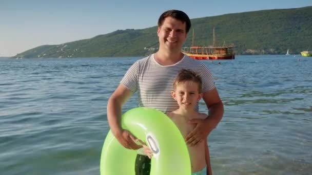 海で泳ぐ前に膨脹可能なリングでビーチに立っている若い父親と幸せな笑顔の少年 家族の休日 夏時間 — ストック動画