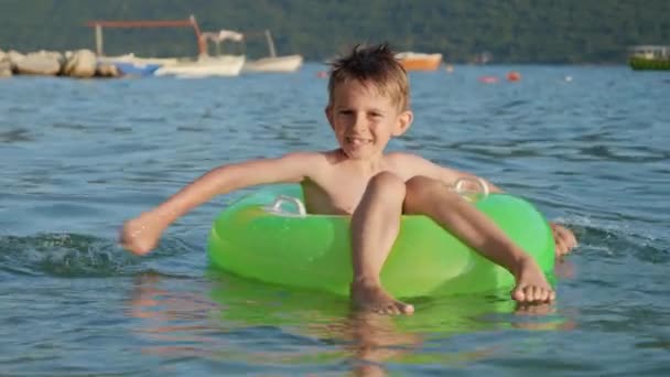 膨脹可能なリングに座り 日没に穏やかな海の波で泳いでいる間 幸せな陽気な少年が水を飛ばします 家族の休日 夏時間 — ストック動画