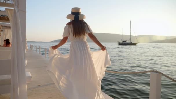 白いドレス 流れる髪の若いブルネットのスローモーションは 海の日没によって木製の桟橋で実行されます 夏時間 ビーチ休暇を引き起こす — ストック動画