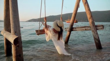 Dalgalı beyaz elbiseli, kumsalda sallanan ve gün batımını seyreden esmer bir kadının yavaş çekim görüntüleri. Turizm, yaz, tatil ve deniz kıyısında tatil..