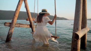 Uzun beyaz elbiseli, kumsalda sallanan ve gün batımını izleyen esmer kadının ağır çekimleri. Turizm, yaz, tatil ve plajda tatil.