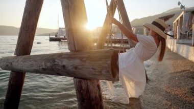 Dalgalı beyaz elbiseli genç esmer kadın, batan güneşin tadını çıkarırken deniz sahilinde ağır çekimde sallanıyor. Seyahat, yaz mutluluğu, tatil rahatlığı ve sahile yakın kaçamaklar..