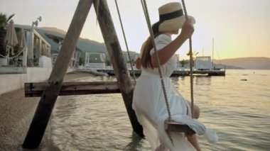 Deniz sahili, zarif, uzun elbiseli bir kadının günbatımında salıncakta sallandığı yer. Turizm, yazın harikası, özgürlüğün neşesi ve seyahat yolculuğu..