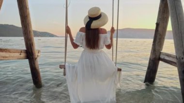 Yavaş çekimde okyanus sahilinde salıncakta sallanan ve deniz suyuna ayaklarıyla dokunan esmer elbiseli genç kadına bakın..