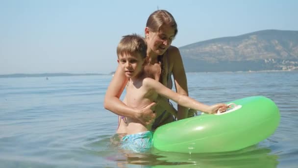 若い世話をする母親は 彼女の小さな息子が海の膨脹可能な水泳リングの中に入るのを助けました 家族の休日 子供と親の楽しい夏時間 — ストック動画