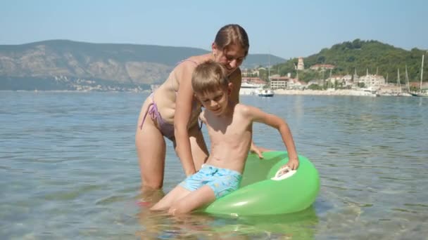 ビキニで幸せな母親は 彼女の息子が膨脹可能な水泳リングに座ってビーチの隣の穏やかな海の波で彼に乗るのを助けます — ストック動画