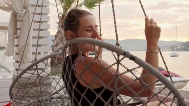 日没の海岸ビーチバーのロープの巣の椅子でリラックスして振動する幸せな笑顔の女性 — ストック動画