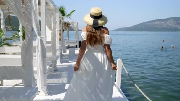 身穿浅色长裙头戴草帽的年轻女子走在海滨的木制码头上 在海上放松的概念 — 图库视频影像