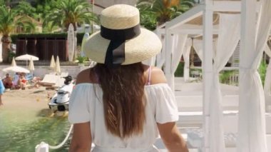 Hafif bir elbise ve hasır şapkayla süslenmiş bir kadın kıyı kaçamağı, deniz kıyısında ahşap bir iskelede yürüyor, tatilin neşesini kucaklıyor..