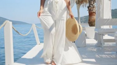 Hafif bir elbise ve hasır şapkayla bir kadının iskelede ağır çekimde yürümesi bayramı, seyahati ve okyanus rahatlığını simgeliyor.