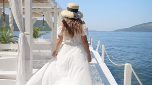 長いドレスのエレガントな女性が木製の海の桟橋を歩き ビーチと海の景色を楽しむガゼボに座る — ストック動画