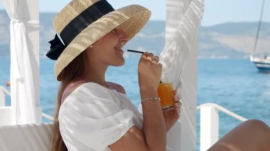 Ağır çekimde hasır şapkalı bir kadın ortaya çıkıyor, ahşap bir deniz iskelesinde bir çardakta. İçkisini yudumlarken, lüks seyahatleri ve yazları sembolize ederek denizde yatları seviyor.