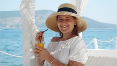 Rıhtımdaki pitoresk bir plaj kafe çardağında neşeli esmer bir kadın oturur ve portakal suyunun tadını çıkarır. Gülüşü yaz, tatil, dinlenme ve seyahatin özünü yansıtıyor..